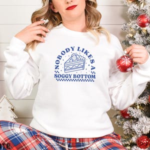 May include: White sweatshirt with blue text that reads "Nobody likes a soggy bottom" and a blue illustration of a slice of cake with a strawberry on top.