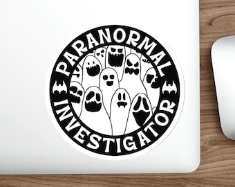 Könnte beinhalten: Ein runder, schwarz-weißer Aufkleber mit dem Text "PARANORMAL INVESTIGATOR", der eine Gruppe von Cartoon-Geistern umkreist. Die Geister haben verschiedene Ausdrücke, einige mit offenem Mund und andere mit einfachen Augen.
