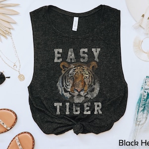 Könnte beinhalten: Schwarzes meliertes Tanktop mit einem Tiger-Grafik und dem Text "EASY TIGER". Das ärmellose Oberteil ist an der Taille geknotet. Sonnenbrillen, Sandalen und Schmuck sind ebenfalls zu sehen.