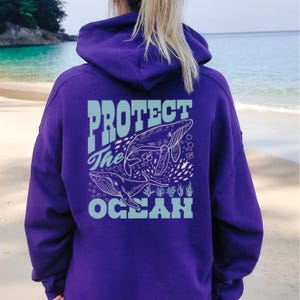 Könnte beinhalten: Lilafarbener Hoodie mit einer weißen Grafik von Walen und dem Text "PROTECT the OCEAN" auf der Rückseite.
