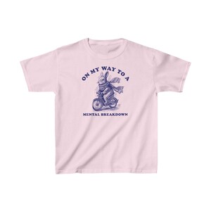 May include: Pink t-shirt featuring a blue illustration of a rabbit riding a bicycle. The text "On my way to a mental breakdown" is printed on the shirt.