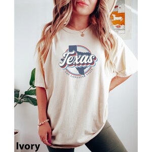 May include: A woman wearing a light ivory t-shirt with a vintage-style graphic of the state of Texas. The graphic says "Texas, The Lone Star State".
