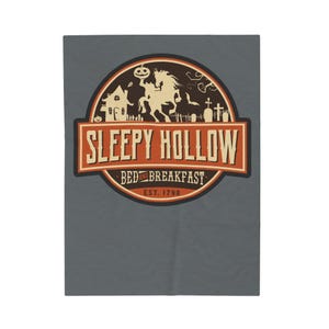 Peut inclure: Une couverture grise avec un motif circulaire portant l'inscription "Sleepy Hollow Bed and Breakfast" en orange et blanc. Le motif comprend un cavalier sans tête, une citrouille, une maison et un cimetière. Un article de décoration intérieure.