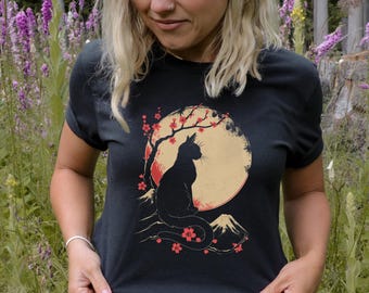 Op de afbeelding: Zwart T-shirt met een afbeelding van een zwarte kat die in een kersenbloesemboom zit voor een volle maan. De maan heeft een rood en gele gradiënt. De kat is gesilhouetteerd tegen de maan.