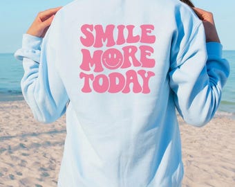 May include: A light blue sweatshirt with a pink retro-style graphic that says "Smile More Today" with a smiley face in the middle of the word "More".