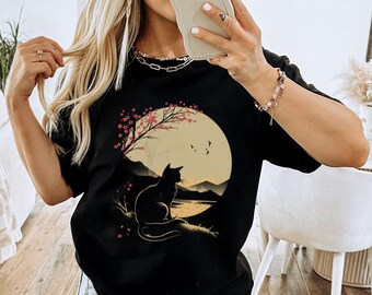 Op de afbeelding: Zwart T-shirt met een afbeelding van een zwarte kat die voor een volle maan met een kersenbloesemboom zit. De maan is geel en de achtergrond is donkerblauw.