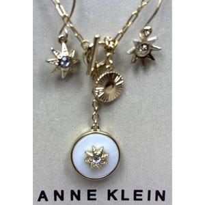 May include: A gold-toned jewelry set featuring a necklace and earrings. The necklace has a white pendant with a starburst design and a small gemstone. The earrings and necklace have star-shaped charms. The brand name "ANNE KLEIN" is printed at the bottom.
