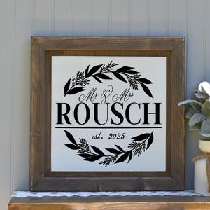 May include: A white wooden sign with a brown frame, featuring a black wreath with leaves and the text "Mr & Mrs ROUSCH est. 2025".