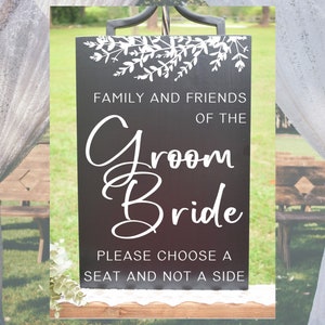 May include: Black wedding sign with white lettering that reads "Family and Friends of the Groom Bride Please Choose a Seat and Not a Side". The sign is decorated with white floral designs.