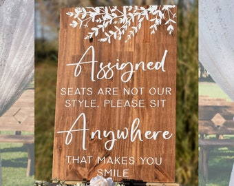 Puede incluir: Un letrero de madera rústica con letras blancas que dice "Assigned Seats are not our style. Please sit anywhere that makes you smile". El letrero está decorado con un diseño floral blanco en la parte superior.