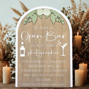 May include: Wooden sign with an arched top, featuring the text "WE HAVE AN Open Bar WE ALSO HAVE A photographer." The sign includes illustrations of a bottle and a martini glass. The sign also includes the text "THIS MEANS ALL DRUNKEN SHENIGANS WILL BE THOROUGHLY DOCUMENTED AND MOCKED FOR YEARS TO COME. *PROCEED AT YOUR OWN RISK.*"