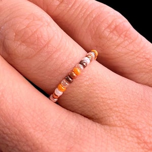 Op de afbeelding: Een delicate kralenring met een mix van oranje, bruine en witte kralen.