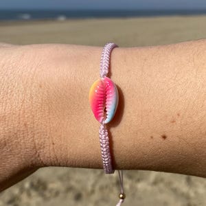 Könnte beinhalten: Ein pinkfarbenes geflochtenes Armband mit einem bunten Muschel-Anhänger. Das Armband ist mit einem Knoten gebunden und hat eine kleine goldene Perle am Ende.
