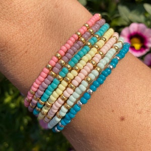 Op de afbeelding: Een stapel kleurrijke kralenarmbanden in tinten roze, geel, blauw en groen. De armbanden zijn gemaakt van kleine, ronde kralen en zijn op elkaar gelegd op iemands pols.