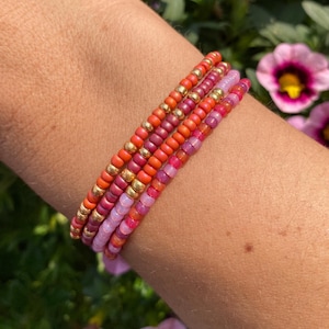 Op de afbeelding: Een stapel van vier kralenarmbanden in tinten roze, rood en goud. De armbanden zijn op elkaar gelegd op de pols van een persoon.
