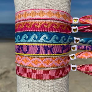 Op de afbeelding: Een stapel kleurrijke geweven armbanden met gouden hartcharms. De armbanden hebben verschillende patronen, waaronder zigzag, golven en ruitjes.
