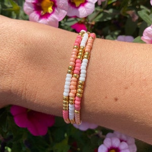 Op de afbeelding: Een set van drie kralenarmbanden in roze, wit en goud. De armbanden zijn gestapeld op de pols van een persoon.