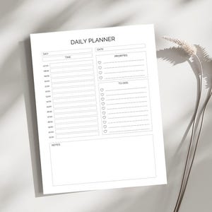 Op de afbeelding: Een wit dagplannerblad met de woorden "DAILY PLANNER" bovenaan. De planner bevat secties voor dag, tijd, datum, prioriteiten en taken. De tijdsectie geeft de uren van 07:00 tot 21:00 uur weer. Een takje gedroogde bloemen staat op de achtergrond.
