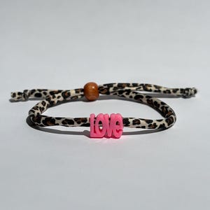 Può includere: Un braccialetto con un cordino a motivo leopardato marrone, nero e beige. Il braccialetto include una singola perla di legno marrone e una perla di plastica rosa brillante a forma della parola love, centrata sul cordino con estremità annodate.