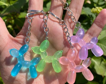 Könnte beinhalten: Vier bunte Ballonhund-Schlüsselanhänger in Blau, Grün, Rosa und Lila. Jeder Hund ist an einer silbernen Kette und einem Ring befestigt. Die Schlüsselanhänger werden in einer Hand vor einem Hintergrund aus grünem Laub gehalten.
