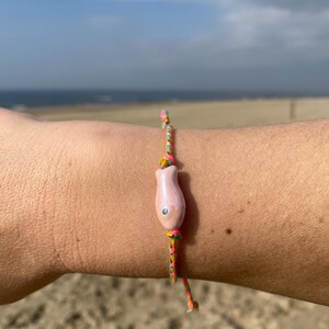 May include: A pink fish charm bracelet with a multi-colored string. The bracelet is on a person's wrist.