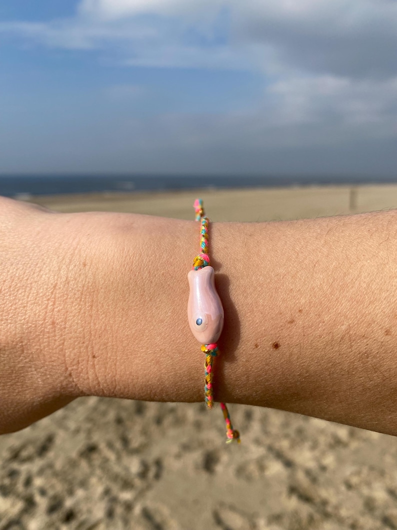 May include: A pink fish charm bracelet with a multi-colored string. The bracelet is on a person's wrist.