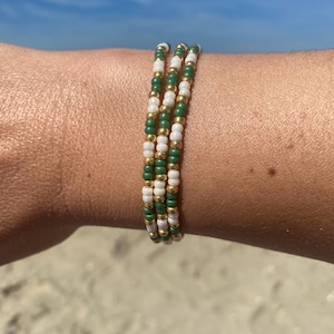 Op de afbeelding: Drie kralenarmbanden met afwisselend witte, groene en gouden kralen. De armbanden worden om een pols gedragen, met een strand en blauwe lucht op de achtergrond. De armbanden zijn gemaakt van kleine, ronde kralen.