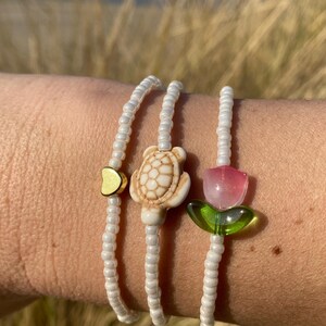 Op de afbeelding: Drie witte kralenarmbanden met bedels. De bedels zijn een gouden hart, een witte schildpad en een roze bloem met groene bladeren.
