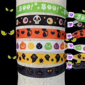 Op de afbeelding: Een stapel Halloween-lintarmbanden. De armbanden hebben verschillende ontwerpen, waaronder geesten, pompoenen, schedels en het woord "BOO!" in het groen. Elke armband heeft een wit koord met een hartvormige kraal.