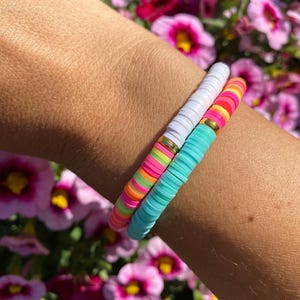 Puede incluir: Dos pulseras de cuentas coloridas en una muñeca. La pulsera superior es blanca con detalles dorados. La pulsera inferior es turquesa, rosa, amarilla y verde.