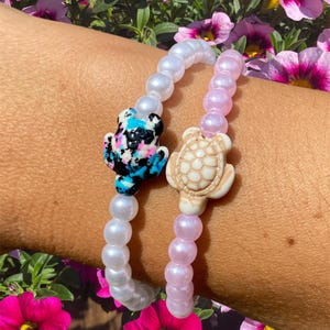 Op de afbeelding: Twee kralenarmbanden met schildpadbedels. De ene armband is wit met een blauwe, zwarte en roze schildpadbedel. De andere armband is roze met een witte schildpadbedel.