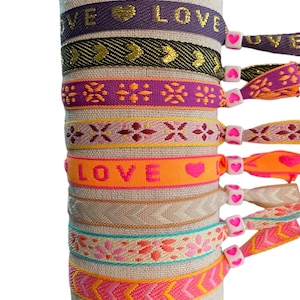 Può includere: Un set di otto braccialetti dell'amicizia colorati con ciondoli a forma di cuore. I braccialetti presentano vari motivi, tra cui disegni geometrici, frecce e la parola "LOVE".