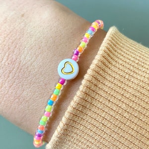 Könnte beinhalten: Ein buntes Perlenarmband mit einem weißen Herz-Anhänger. Die Perlen sind regenbogenfarben und der Anhänger hat einen goldenen Umriss.