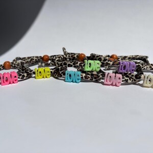Könnte beinhalten: Eine Sammlung von Armbändern mit einem Stoffband im Leopardenmuster, einer kleinen braunen Holzperle und einem zentralen Kunststoffanhänger in Form des Wortes love. Die Anhänger sind in verschiedenen Farben erhältlich, darunter Pink, Gelb, Blau, Limettengrün, Hellrosa, Lila und Weiß.