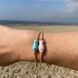 May include: Two colorful bracelets with fish charms. The bracelets are made of thin, braided cords in shades of yellow, pink, and blue. The fish charms are made of ceramic and have a blue and pink glaze. The bracelets are worn on a person's wrist.