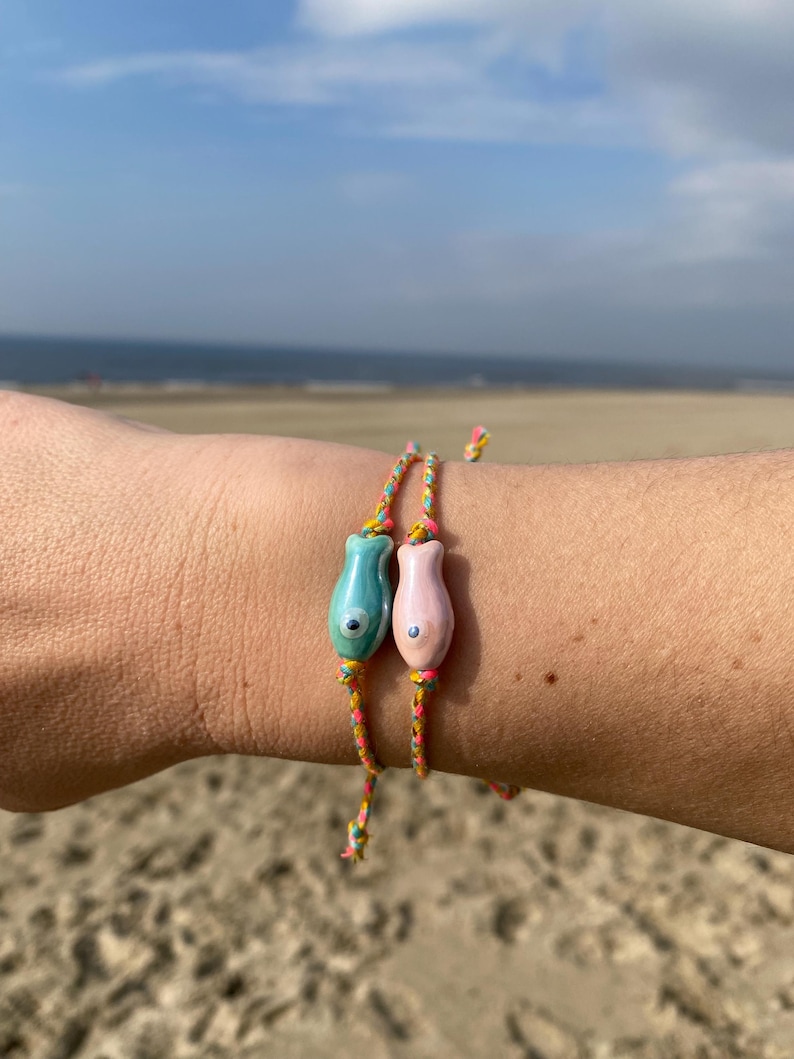 May include: Two colorful bracelets with fish charms. The bracelets are made of thin, braided cords in shades of yellow, pink, and blue. The fish charms are made of ceramic and have a blue and pink glaze. The bracelets are worn on a person's wrist.