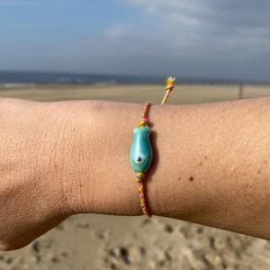 May include: A colorful string bracelet with a turquoise blue and white evil eye charm. The bracelet is worn on a person's wrist against a sandy beach background.