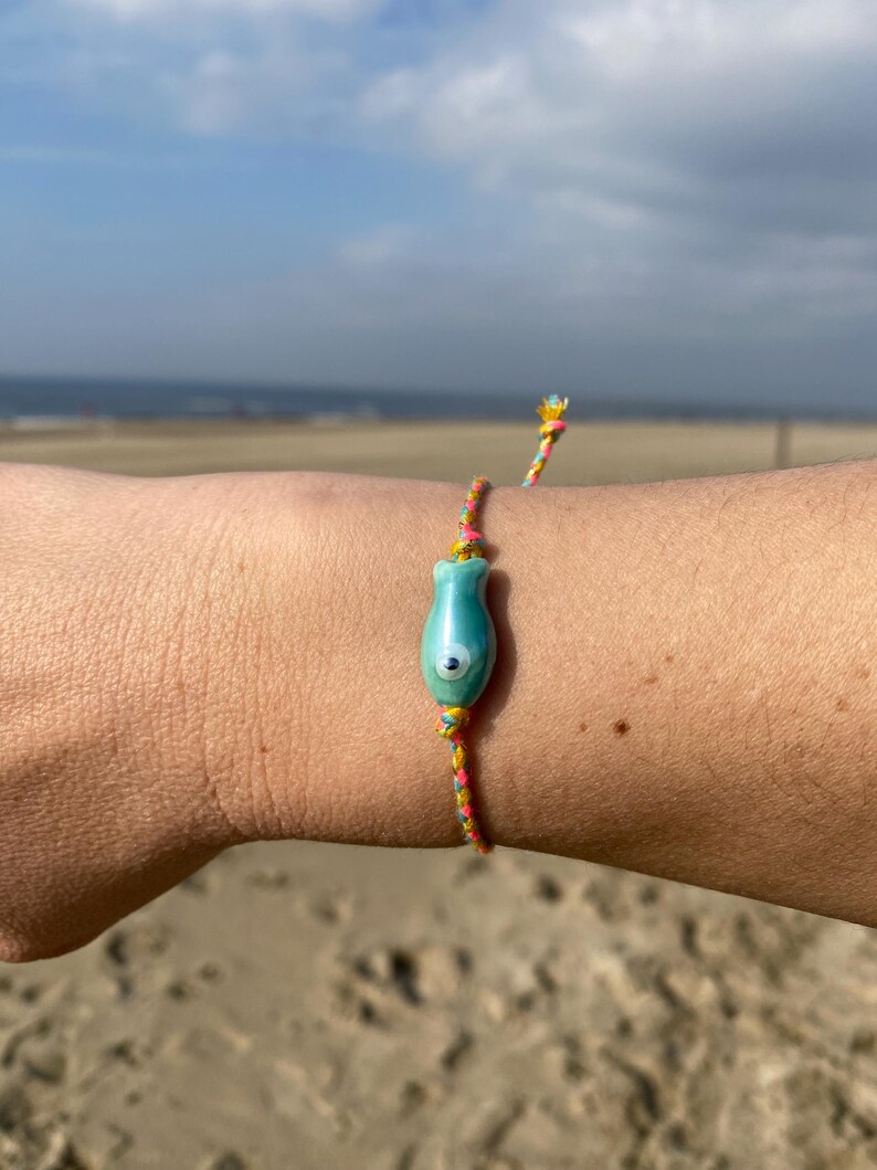 May include: A colorful string bracelet with a turquoise blue and white evil eye charm. The bracelet is worn on a person's wrist against a sandy beach background.