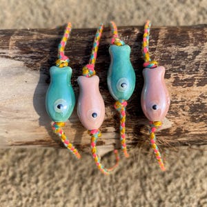 Op de afbeelding: Vier kleurrijke armbanden met visvormige bedels. De bedels zijn gemaakt van keramiek en hebben een blauwe en roze glazuur. De armbanden zijn gemaakt van gevlochten koord in geel, roze en blauw.