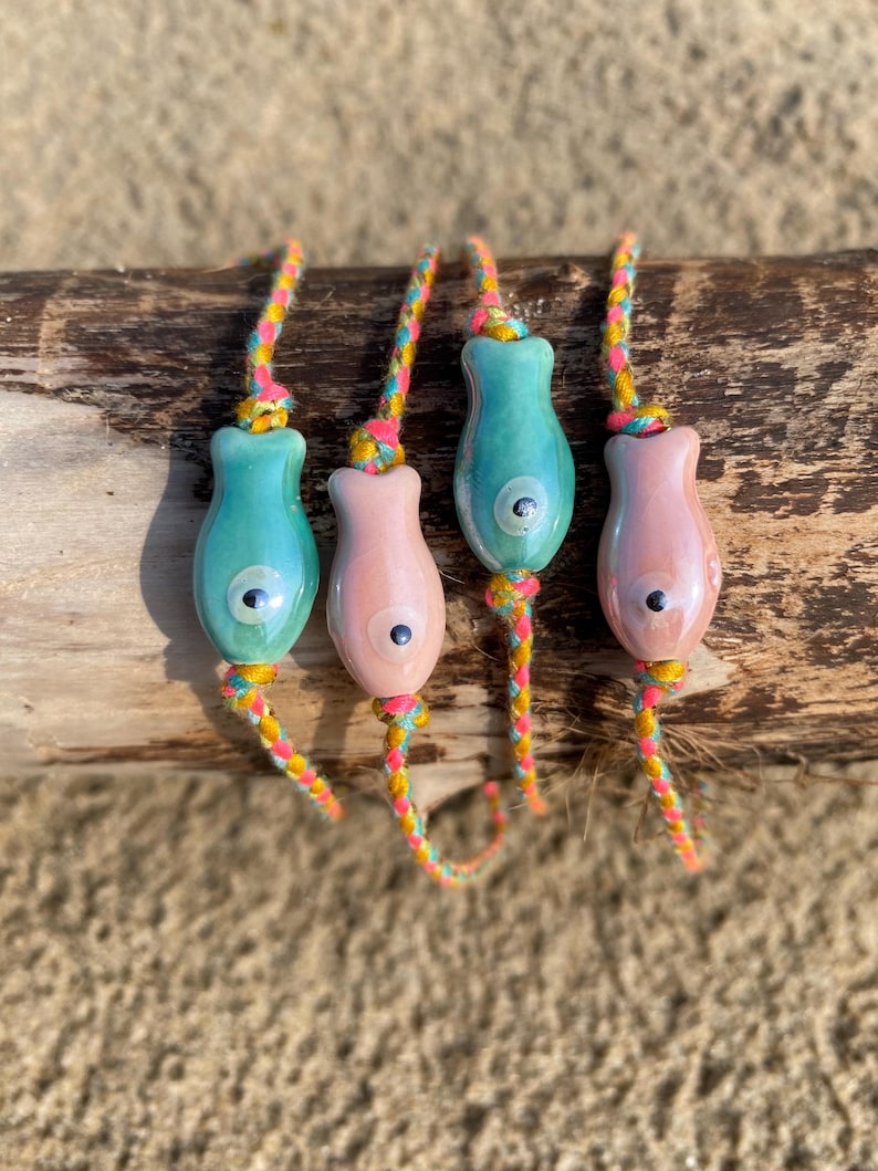 May include: Four colorful bracelets with fish-shaped charms. The charms are made of ceramic and have a blue and pink glaze. The bracelets are made of braided cord in yellow, pink, and blue.