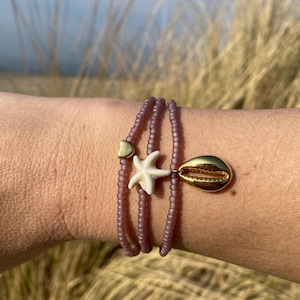 Op de afbeelding: Een gelaagde armband met paarse kralen en gouden accenten. De armband heeft een klein hartje, een zeester en een schelp.