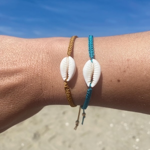 Puede incluir: Dos pulseras con un dije de concha de cauri. Una pulsera es azul turquesa y la otra es marrón.