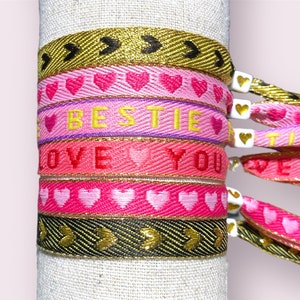 Op de afbeelding: Een set van vijf vriendschapsarmbanden met verschillende ontwerpen. De armbanden zijn gemaakt van geweven stof in de kleuren roze, rood, goud en zwart. De armbanden bevatten harten, het woord "BESTIE" en de woorden "LOVE YOU".