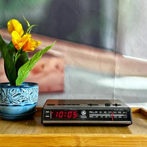 Puede incluir: Un radio reloj despertador digital General Electric con acabado de imitación madera y panel frontal negro. La pantalla LED roja muestra la hora 10:05. La radio cuenta con un dial de sintonización analógico para frecuencias AM y FM.