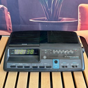 Puede incluir: Radio reloj estéreo AM/FM GE vintage. La pantalla digital muestra la hora en verde. Incluye función de repetición, conector para auriculares y dos configuraciones de despertador. La radio tiene un diseño negro y gris.