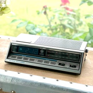 Może przedstawiać: Srebrny radiobudzik General Electric z elektronicznym sterowaniem dotykowym, cyfrowym wyświetlaczem pokazującym 2:12, analogowym tunerem FM/AM oraz dwoma przełącznikami czasu budzenia. Urządzenie posiada górną kratkę głośnika i przyciski.