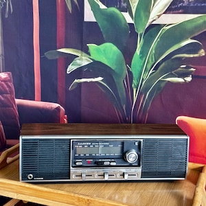 Könnte beinhalten: Ein rechteckiges AM/FM-Stereoradio der Marke Lloyd's mit einer Oberseite in dunkler Holzmaserung und schwarzen Lautsprechergittern an den Seiten. Die Frontblende verfügt über ein zentrales Abstimmrad, einen Kopfhöreranschluss und drei silberne Regler.