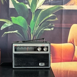 Könnte beinhalten: Ein Vintage Hitachi Transistorradio in Schwarz und Silber. Das Radio hat ein Einstellrad, Lautstärkeregler und einen Tragegriff. Auf der Vorderseite stehen die Worte "AC-BATTERY" und "SOLID STATE".