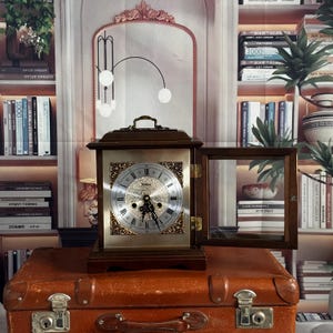 Peut inclure: Une horloge en bois de style vintage avec un cadran doré et des chiffres romains. L'horloge est rectangulaire, avec une poignée et une porte vitrée. Elle est posée sur une valise marron, devant une bibliothèque.