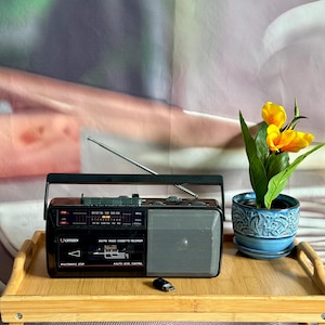 May include: A black Citizen AM/FM radio cassette recorder with a silver speaker grille, a retractable antenna, and a black carrying handle. The device features a cassette deck, radio tuning dial, and a small black USB drive placed in front.